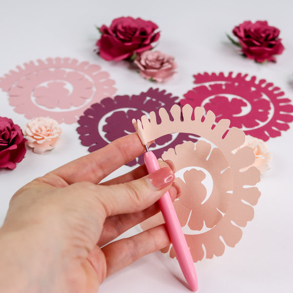 Quilling tool rolling up a pink rolled rose paper flower with paper rolled roses in background.
